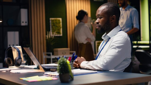 Black man accountant in office using laptop and writing down information, working late to finish tasks before deadline, transcribing from screen and taking notes, documenting research.