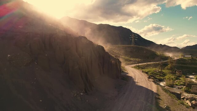 Aerial tracking shot of winding dirt road along rocky sand cliffs and foothills in Altai mountainous countryside with sun flare.