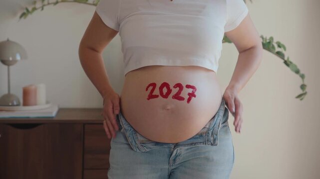 4K video of a pregnant woman with the red number 2027 painted on her bare belly, symbolizing her baby's due date year and the countdown to motherhood