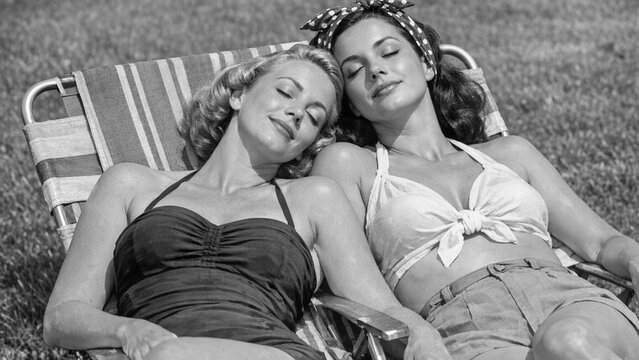 Two beautiful women enjoying a lazy day at the beach. They are dressed in vintage swimwear and seem to be having a great time together.