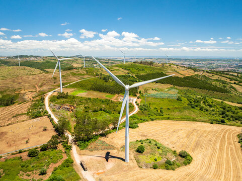 Aerial view of wind turbines standing in the rolling hills of the countryside with green vegetation and dry fields under a bright blue sky in Sintra, Lisbon, Portugal.
