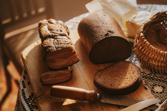 Detailed close-up view of a rustic wooden cutting board featuring a large piece of smoked pork bacon (speķis) being sliced next to a traditional loaf of dark rye bread and a vintage kitchen knife.