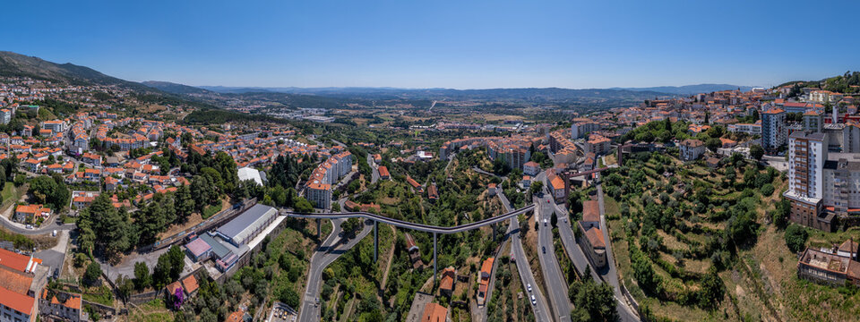 Aerial view of the Carpinteira Pedestrian Bridge spanning the valley with urban buildings, winding roads, and lush green hillsides in the city of Covilha, Castelo Branco District, Portugal.