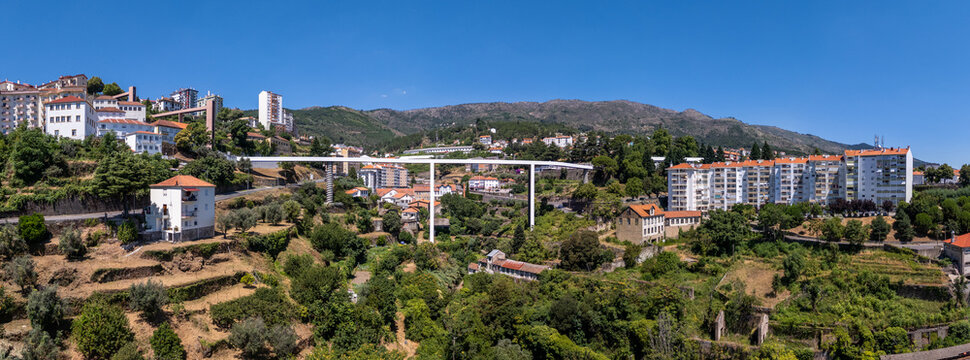 Aerial view of Carpinteira Pedestrian Bridge spanning the valley with modern residential buildings, lush green hillsides, and clear blue sky in Covilha, Castelo Branco District, Portugal.