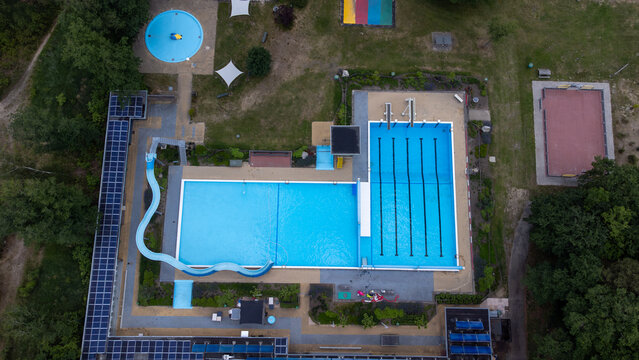 Aerial view of the swimming pool at Beleefpark Dwingeloo featuring a blue basin with swimming lanes, a water slide, and solar panels on the building roof in Dwingeloo, Drenthe, Netherlands.