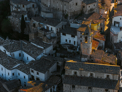 Aerial view of the historic town of Capestrano with stone buildings, terracotta rooftops, and a church bell tower illuminated by warm golden hour light in Capestrano, Abruzzo, Italy.