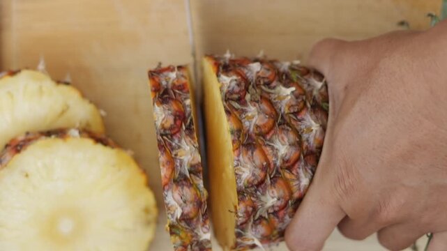 Close-up hand slicing fresh pineapple on wooden cutting board with knife. Tropical fruit preparation concept showing healthy food, natural ingredients and fresh organic pineapple texture in kitchen.