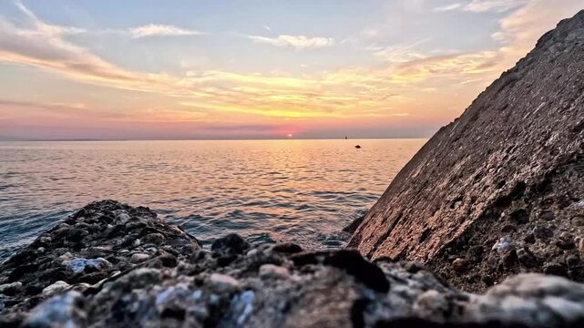 Time-lapse sunset over the Mediterranean Sea framed by rocks as two sailboats move on the horizon. The video is part of a series filmed during a seaside holiday in the seaside village of Rodia, Sicily