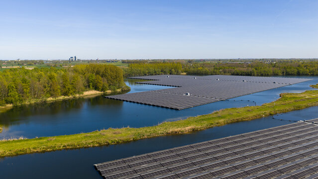 Aerial view of a large floating solar farm on a calm lake surrounded by lush green trees and fields under a clear blue sky in Zwolle, Overijssel, Netherlands.