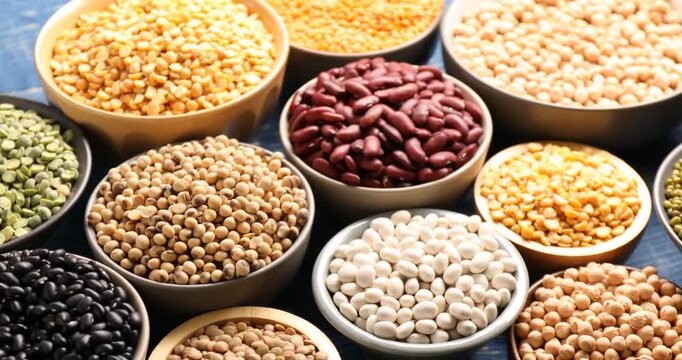 Different types of legumes in bowls on blue wooden table, spinning. Closeup view