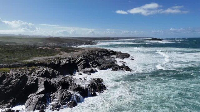 Aerial drone shot pushing in along the stunning, rugged coastline of Portugal, showcasing powerful waves crashing against cliffs and rocky shores under a bright blue sky. Part of the Rota Vicentina.