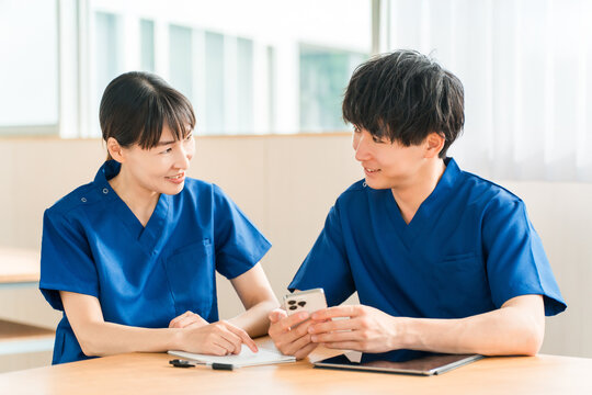 A senior nurse and a young male nurse are talking while looking at their smartphones.