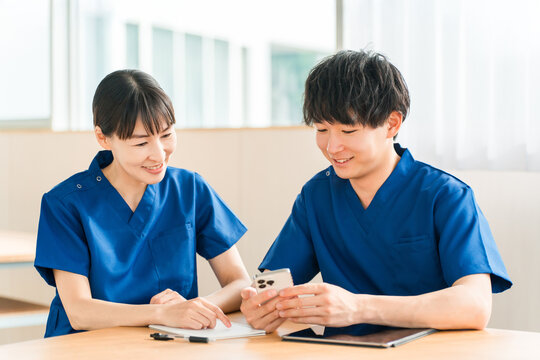 A senior nurse and a young male nurse are talking while looking at their smartphones.
