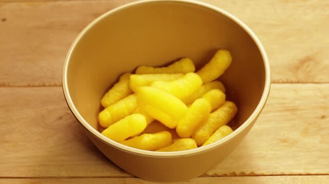 Yellow corn sticks being poured into a brown bowl on a rustic wooden table. Processed corn snack.