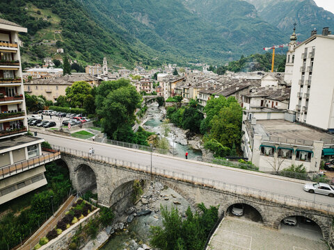 Aerial view of the historic town of Chiavenna, featuring a stone arch bridge over the Mera River, surrounded by lush green mountains and traditional buildings in Chiavenna, Lombardy, Italy.