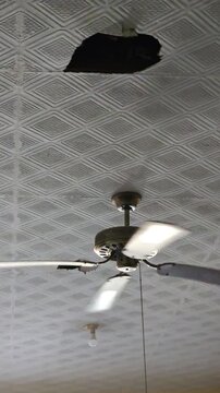 A spinning ceiling fan installed below a patterned ceiling featuring a noticeable damaged hole in the tiles
