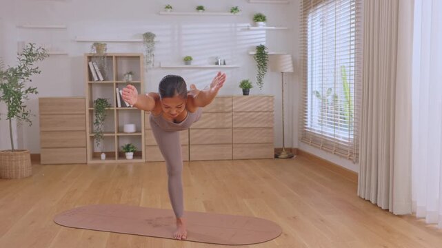 Determined Asian woman wearing purple activewear, practicing calm yoga balance pose at home, executing healthy fitness exercise on mat for strength, serenity during morning training session