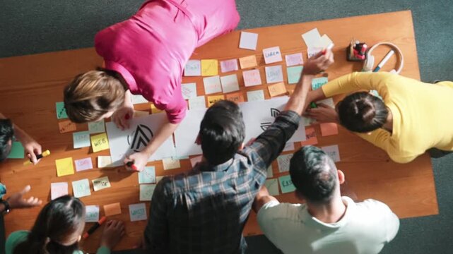 Top down aerial view of skilled designer hold logo while discuss with team. Aerial business group working together sharing idea about logo graphic design by using sticky notes and tablet. Symposium.