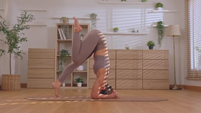Determined athletic woman performs challenging headstand yoga pose at home, showcasing physical strength, balance, flexibility during daily fitness exercise, peaceful mind workout session
