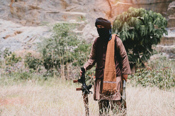 Fototapeta premium Outdoor military concept. Masked armed man in dry grass near rocky cliff, rural conflict scene