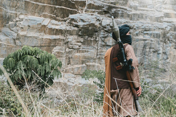 Fototapeta premium Outdoor military concept. Desert warrior with rocket launcher stands near rocky cliff in dry grass