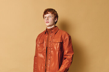 Obraz premium Young man with red hair wearing orange leather jacket and turtleneck stands against beige background looking forward with calm serious expression.