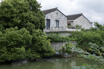 Fototapeta premium Weathered Jiangnan whitewashed house beside a calm river surrounded by lush vegetation in rural Zhejiang China. Tranquil riverside dwelling, cultural heritage, rural homes, traditional lifestyle
