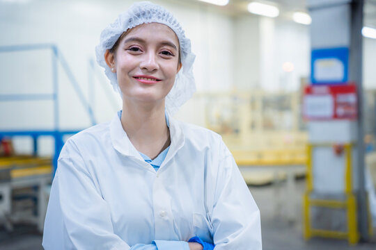 Smiling female quality control technician in white lab coat and protective hair net at work, Portrait of a confident female food factory worker standing in a clean production plant