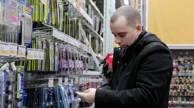 Focused hardware store customer carefully examining drill bits on display rack, critically evaluating tool options before making strategic purchasing decision