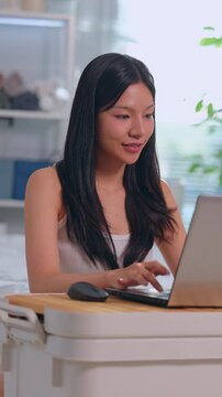 Happy young asian adult woman smiling while working on laptop computer in bright home office, typing on desk with mouse, enjoying remote work, online learning, positive emotional expression Vertical