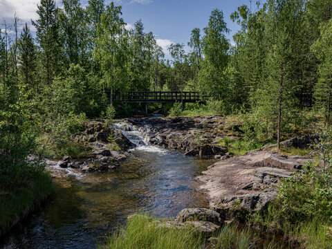 Small rapids and a wooden footbridge at the Storforsen Nature Reserve on the Pite River in &Auml;lvsbyn Municipality, Norrbotten, Sweden. Crystal-clear water flowing through rocky terrain surrounded by lus