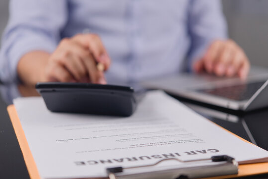 Close up of a car insurance policy document on a clipboard with a blurred person calculating premiums in the background, symbolizing vehicle protection, liability coverage, and auto risk control.