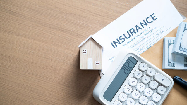 Hand calculating 2027 home insurance expenses beside cash bundles, miniature house, policy document, envelope, and pen on a wooden table.