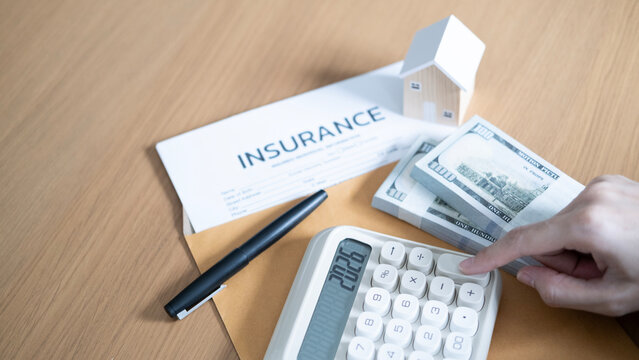 Home insurance and mortgage cost calculation with a hand using calculator, wooden house model, cash bundles, policy form, envelope, and pen.
