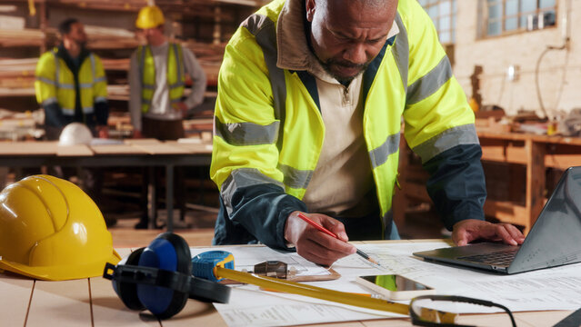 Laptop, man and drawing in warehouse for blueprint, manufacturing and woodwork contractor. Paperwork, production and carpenter person in joinery factory with tech in workshop, draft or project