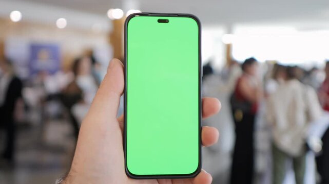 Hand holds smartphone with green screen. Blurred crowd moves in background. Useful for app demos or digital content. Focus on phone. Ideal for UI design, presentations, or tech visuals