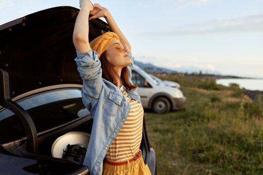 Cheerful woman outdoors, stretching with arms raised near an open trunk, sunlit countryside, striped top, denim jacket, carefree mood, natural light, relaxed lifestyle and breeze