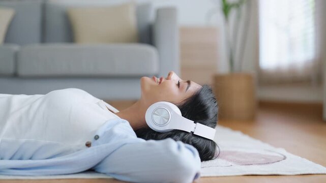 Young Asian woman relaxing at home. Girl in headphones listen to music in cozy living room, using audio healing apps for mental health. Female wellness and self-care lifestyle, ASMR sound therapy