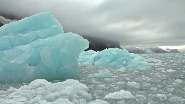 Magnificent turquoise ice structures drift gracefully across chilly Arctic waters of Svalbard, Norway. This serene summer scene showcases frozen beauty distant, mist-shrouded peaks under cloudy sky.