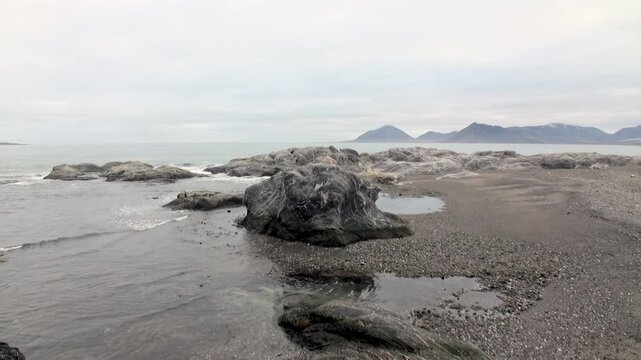 Gentle waves ebb and flow across a pebbled beach in Svalbard, Arctic Norway. Rugged rocks dot the shoreline under a vast, overcast sky.