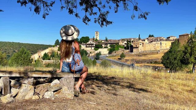 Woman with hat on bench looking at medieval village of Pedraza Spain