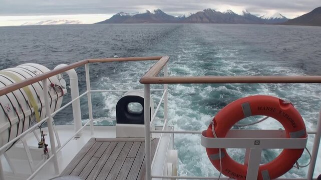 Vessel charts course through open Arctic sea near Svalbard, Norway. Churning wake paints frothy path behind ship under muted summer sky, with distant snowy mountains gracing horizon.