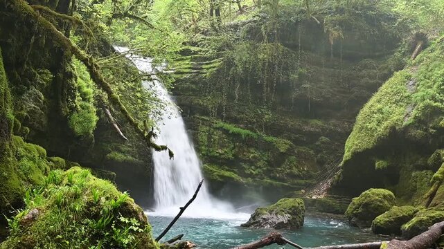 A powerful waterfall plunging into a clear turquoise pool surrounded by a deep gorge with moss-covered rocks and lush green vegetation
