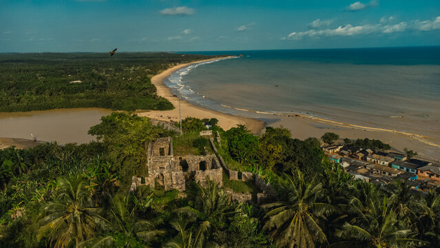 Aerial view of the historic ruins of Fort Batenstein surrounded by lush palm trees, overlooking the sandy beach and the river mouth in Butre, Western Region, Ghana.