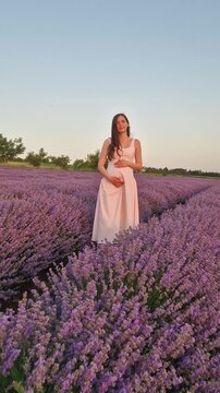 A beautiful pregnant woman smiles and poses in a vast lavender field. Wearing a pink dress and straw hat, she cradles her baby bump, enjoying the endless rows of purple lavandula flowers at sunset