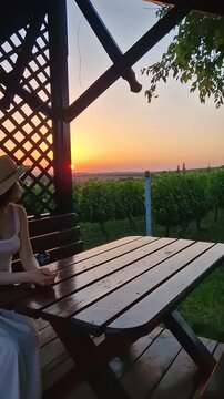 A beautiful woman sits inside a wooden gazebo overlooking a scenic vineyard at sunset. Wearing straw hat and light dress, she relaxes at a table, admiring the rows of grapevines under the setting sun