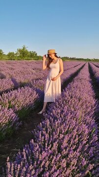 A beautiful pregnant woman smiles and poses in a vast lavender field. Wearing a pink dress and straw hat, she cradles her baby bump, enjoying the endless rows of purple lavandula flowers at sunset