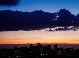 Obraz premium City skyline silhouette at dusk. Dark clouds cover the orange and blue twilight sky. Building lights twinkle in the darkening urban landscape.