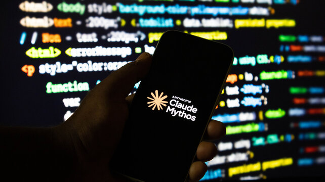 Hand holding a smartphone displaying the anthropic claude mythos logo in front of a computer screen with programming code