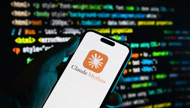 Closeup of a smartphone displaying the claude mythos logo held in front of a computer screen with programming code